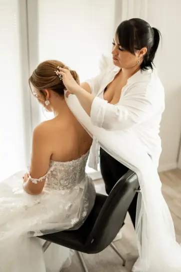 Eine Make-up Visagistin befestigt einen Schleier mit einer perlenbesetzten Haarspange an der Hochsteckfrisur einer Braut, die in einem eleganten, Hochzeitskleid mit Spitzenverzierungen sitzt.