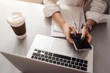 Webdesignerin die am Pc sitzt und ein Handy hält, daneben steht eine Kaffeebecher.